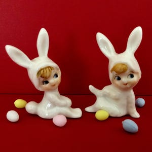 May include: Two ceramic figurines of children in white bunny suits with painted faces and blue eyes. Colorful Easter eggs surround the figurines on a red background. A charming Easter decoration.