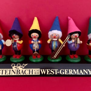 May include: A collection of Steinbach West-Germany wooden gnome figurines, each playing a different musical instrument. The gnomes wear colorful outfits and hats, including red, purple, yellow, blue, pink, and green. The base is black with gold lettering.