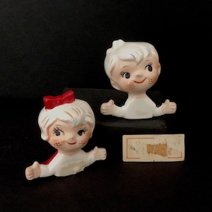 Puede incluir: Un par de figuritas de cerámica vintage de niños con piel blanca, cabello rubio y detalles rojos. La niña tiene un lazo rojo en la cabeza y el niño tiene una camisa verde. Las figuritas están en el estilo de un dibujo infantil.