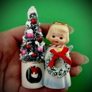 Könnte beinhalten: Vintage Weihnachtsfigur mit einem blonden Engel mit goldenem Heiligenschein und roter Schleife, der einen verschneiten Kranz hält. Daneben ein Miniatur-Weihnachtsbaum mit roten und rosa Ornamenten und einem weißen Sockel mit grünem Kranzdesign.