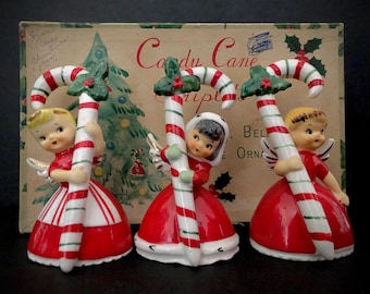 Może przedstawiać: Trzy figurki aniołów bożonarodzeniowych w stylu vintage trzymające ozdoby w kształcie lasek cukrowych. Anioły mają na sobie czerwone sukienki z białymi wykończeniami i złote skrzydła. Laski cukrowe są w czerwono-białe paski z zielonymi akcentami ostrokrzewu. Tło przedstawia pudełko ze słowami "Candy Cane Scripts".