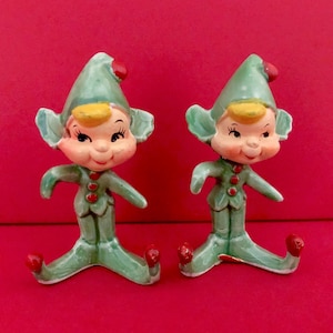 Könnte beinhalten: Zwei grüne Weihnachtswichtel aus Keramik mit roten Mützen und gelbem Haar. Die Wichtel stehen auf einer roten Oberfläche.