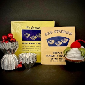 May include: A set of silver-colored fluted metal baking molds with a yellow box labeled Old Swedish Ebba's Forms & Recipes, a tan booklet titled Old Swedish Ebba's Forms & Recipes Estab. 1954, and a small decorative cupcake ornament with a red cherry.