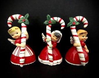 May include: Three ceramic Christmas angel figurines. Each angel wears a red dress with white stripes and holds a candy cane. The candy canes are red and white striped with green holly accents. The angels have blonde or brown hair and small wings.