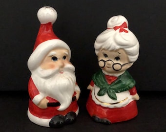Könnte beinhalten: Ein vintage Keramik-Salz- und Pfefferstreuer-Set mit Weihnachtsmann und Frau Claus. Der Weihnachtsmann trägt einen roten Anzug mit weißem Bart und einem schwarzen Gürtel. Frau Claus trägt ein rotes Kleid mit einem grünen Schal und einer weißen Schürze. Beide Figuren haben bemalte Details und sind in gutem Zustand.