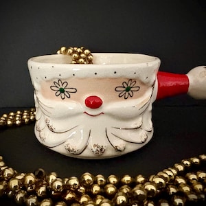 May include: A ceramic Santa Claus face-shaped bowl with a red handle. The bowl is white with a pink face, black eyes, and a red nose. Gold beads are inside the bowl and around it. The bowl has a vintage, festive design.