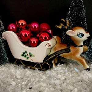 May include: A festive Christmas scene featuring a ceramic reindeer pulling a white sleigh filled with red ornaments. The sleigh is decorated with a green floral design. Miniature Christmas trees and faux snow complete the holiday display.