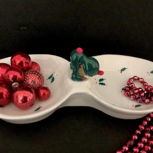 Könnte beinhalten: Weiße, geteilte Keramikschale mit einer grünen und roten Weihnachtsbaumfigur. Die Schale hat ein Design mit Stechpalmenblättern und ist mit roten Weihnachtskugeln und rosa Perlen gefüllt.