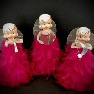 May include: Three vintage Christmas angel figurines with pink feather skirts and clear plastic wings. Each angel holds a musical instrument: a flute, a book, and a crescent moon.