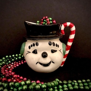 May include: White ceramic snowman creamer with a black top hat and a candy cane handle. The creamer is filled with red and green Christmas beads.