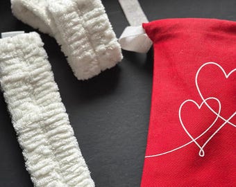 Puede incluir: Un conjunto de diademas de spa blancas y esponjosas y una bolsa roja con cordón y diseño de corazón blanco. Las diademas son rectangulares con una superficie texturizada y absorbente. La bolsa es de tela lisa.