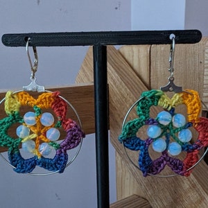 May include: Rainbow crochet earrings with iridescent beads and silver hooks. The earrings are displayed on a black stand.