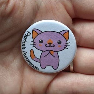 Puede incluir: Un botón redondo con una ilustración de gato de dibujos animados en morado y naranja. El botón tiene el texto "Psoriasis Awareness" en una fuente curva alrededor del gato. El botón se sostiene en una mano.