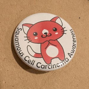 May include: A round button with a red cartoon cat illustration. The cat is standing with its paws together. The button has the text "Squamous Cell Carcinoma Awareness" around the edge.