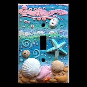 May include: Decorative light switch cover featuring a beach scene. The cover is painted in shades of blue, white, and tan, with sculpted seashells, a starfish, and other sea-themed elements. The switch opening is black.