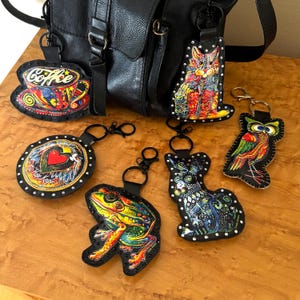 May include: A collection of colorful leather keychains featuring various designs, including a coffee cup, a heart, a cat, an owl, a frog, and a rabbit. Each keychain has a black border with silver studs and a metal clasp.