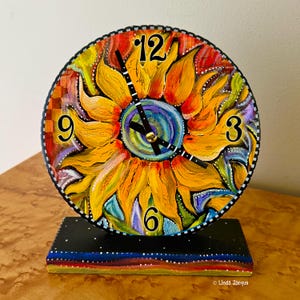 May include: A hand-painted, round clock featuring a vibrant sunflower design. The clock face displays the numbers 3, 6, 9, and 12 in black. The clock is set on a black rectangular base with a colorful, striped bottom. The artist's signature is visible.