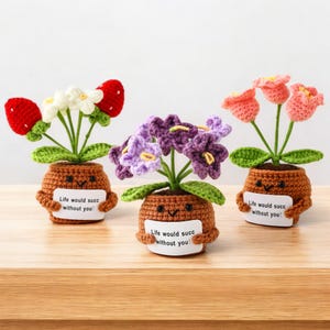 May include: Three crocheted potted plants with brown yarn pots featuring embroidered faces and arms holding white signs that read "Life would succ without you!". The plants include red strawberries with white flowers, purple flowers, and pink tulip-like flowers.
