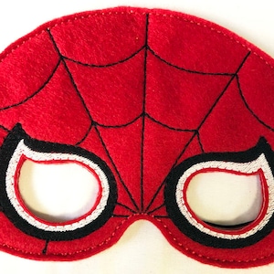 May include: A red felt mask featuring a black embroidered spiderweb pattern across the surface. The mask has two eye openings outlined with thick black and white stitching. A thin black elastic cord is attached to each side of the mask.