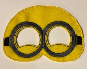 May include: A yellow felt eye mask with two black eye holes. The eye holes are outlined in black stitching.