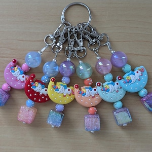 May include: A collection of keychains featuring colorful unicorn designs on crescent moons. Each keychain has a silver clasp, iridescent beads, and a square, glittery charm. The keychains are in shades of pink, red, yellow, orange, blue, and purple.