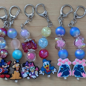 May include: Keychains with silver-tone hardware. Each keychain features a series of colorful beads and a character charm. Charms include Mickey Mouse, Pluto, Minnie and Daisy Duck, Donald Duck, and Stitch.