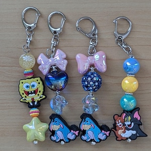 May include: Four keychains with cartoon character charms. One features SpongeBob SquarePants, a yellow star, and colorful beads. Another has a pink bow and a blue heart. The third has a pink bow and a blue beaded ball. The last has a blue, yellow, and teal bead design with Tom and Jerry.