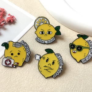 May include: Four yellow lemon-shaped enamel pins with black outlines and green leaves. Each pin has a different facial expression and text: "Lemon-Aid", "bitter lemon", "A little bitter", and "Live life with zest".