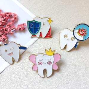 May include: Set of four enamel pins featuring cartoon teeth. One tooth has a toothbrush, another a shield, a third a magnifying glass with germs, and the last a crown and pink wings. Each pin has gold-colored trim.