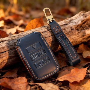 May include: A dark navy blue leather car key fob cover with embossed button icons and contrasting orange stitching, accompanied by a matching leather keychain strap featuring a brass-toned metal lobster clasp and a square stitched detail.