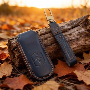 May include: A dark blue leather key fob cover with embossed lock and unlock icons, paired with a matching leather strap featuring a gold-toned metal swivel clasp. The items are stitched with thick brown thread along the edges.