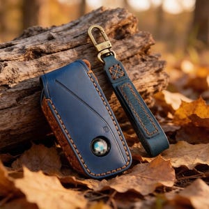 Peut inclure: Un étui à clé en cuir bleu marine avec des coutures marron et un emblème circulaire BMW, accompagné d'une lanière en cuir bleu marine assortie dotée d'un mousqueton en métal couleur laiton et d'une étiquette en cuir avec du texte gaufré.