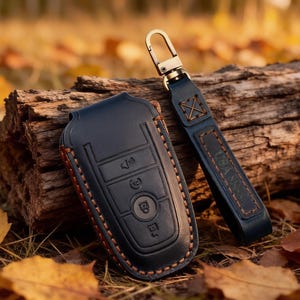 May include: A dark navy blue leather car key fob cover featuring embossed button icons and contrasting tan stitching. The cover is attached to a matching leather strap with a gold-toned metal lobster clasp, showcasing a structured design.