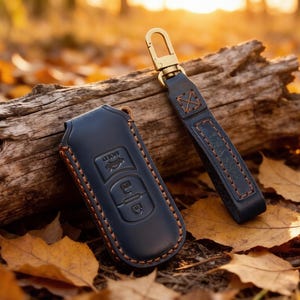 May include: A dark blue leather key fob cover with visible orange stitching and a matching leather strap featuring a gold-toned metal swivel clasp. The cover has embossed icons for lock, unlock, and boot release functions on its front surface.