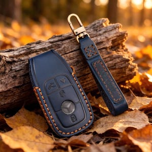 May include: A dark blue leather car key fob cover with visible orange stitching and a matching leather keychain strap featuring a gold-toned metal clasp. The key cover has embossed button icons and a circular centre cutout for a vehicle emblem.