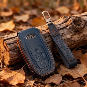May include: A dark blue leather key fob cover for an Audi vehicle, featuring contrasting orange stitching and a matching leather strap with a metal lobster clasp. The cover has embossed button icons and a cutout for the Audi logo emblem.