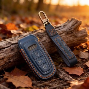 May include: A dark blue leather car key fob cover with orange stitching and a matching leather keychain strap featuring a metal clasp. The key fob cover has three embossed buttons and a small cutout, while the strap displays embossed text.