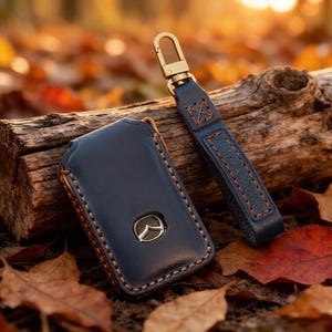 May include: A dark blue leather car key fob cover featuring contrasting tan stitching and a metallic Mazda logo emblem. The cover is attached to a matching leather strap with a brass-toned metal lobster clasp hook for secure attachment.