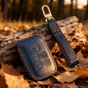 May include: A dark blue leather key fob cover with embossed button icons and a matching leather strap featuring a gold-toned metal lobster clasp. The cover and strap are stitched with thick brown thread, showcasing a durable construction.