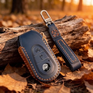 May include: A dark blue leather car key fob cover with contrasting orange stitching and a matching leather keychain strap featuring a metal swivel clip. The key cover has moulded button icons and a circular cutout for the emblem.