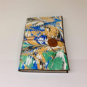 May include: A rectangular journal featuring a marbled paper cover with a vibrant pattern of blue, gold, green, black, and white swirls. The journal is secured with a thin purple cord wrapped around a circular brown button and a small green bead.
