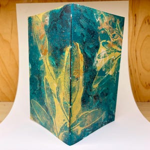 May include: A folded card featuring a deep teal and dark blue textured background with a prominent yellow botanical leaf print. The design displays a layered, painterly aesthetic with visible brushstrokes and a vertical crease down the center.