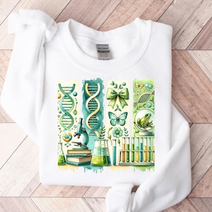 Puede incluir: Sudadera blanca con un diseño gráfico colorido que presenta un microscopio, hebras de ADN, tubos de ensayo, una rana, una mariposa y un lazo. El diseño es de estilo acuarela con colores verde, azul y amarillo.