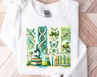May include: White sweatshirt with a colorful graphic design featuring a microscope, DNA strands, test tubes, a frog, a butterfly, and a bow. The design is in a watercolor style with green, blue, and yellow colors.