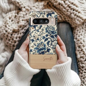Pode incluir: Uma capa de telefone com um design floral em tons de azul e creme. A parte superior apresenta um padr&atilde;o detalhado de flores e folhas, enquanto a parte inferior tem um acabamento em gr&atilde;o de madeira com o nome "Samantha" em escrita cursiva.