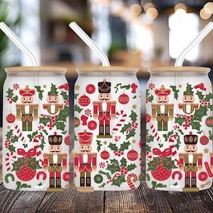 Könnte beinhalten: Drei Glasbecher mit Bambusdeckeln und klaren Strohhalmen. Jeder Becher hat ein festliches Weihnachtsdesign mit Nussknackern, Stechpalmen, Zuckerstangen und Ornamenten. Der Hintergrund ist weiß.