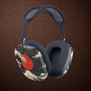 May include: Over-ear headphones featuring a dark blue headband and ear cushions. The outer ear cups display a graphic print of a large red sun rising behind stylized crashing waves and mountain peaks in shades of black, white, and cream.
