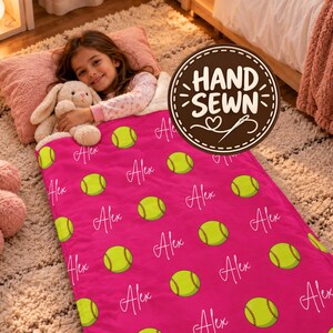 May include: A bright pink blanket featuring a repeating pattern of yellow softballs and the name Alex written in white cursive script. The blanket has a soft texture and is displayed with a plush toy.