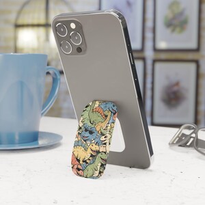 Könnte beinhalten: Ein Smartphone mit einem farbenfrohen Handyhalter mit Dinosaurier-Motiv. Der Halter zeigt verschiedene Cartoon-Dinosaurier in Grün-, Blau-, Gelb- und Orangetönen. Das Telefon ist auf einer weißen Oberfläche aufgestellt, was eine freihändige Nutzung ermöglicht.