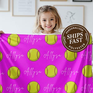May include: A personalized pink softball blanket featuring a pattern of yellow softballs and the name "Allysa" in white script. The blanket is made of polyester and has a "Ships Fast" logo.
